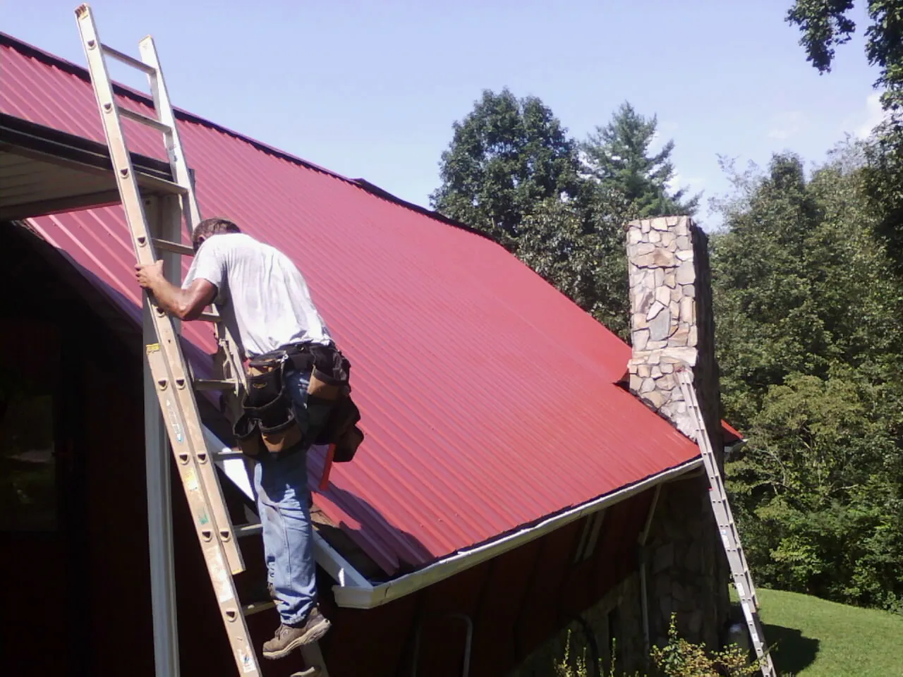 Roofer on a ladder beside a red metal roof and stone chimney — Soffit & Fascia Repair by Roofing Brothers in Erie